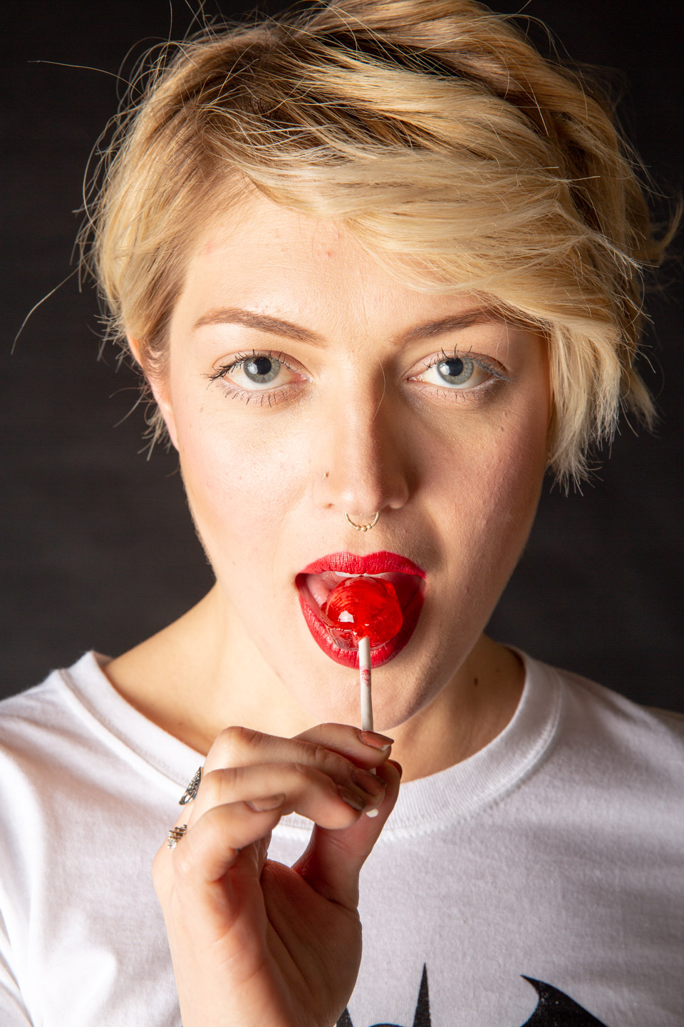 Woman with short blonde hair holding red lollipop against dark background
